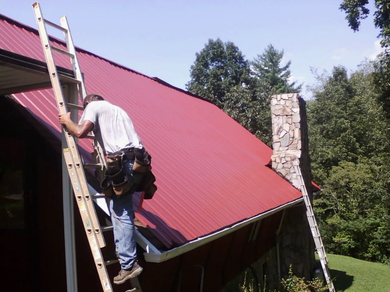 Roofer on a ladder beside a red metal roof and stone chimney — Soffit & Fascia Repair by Roofing Brothers in Okeana